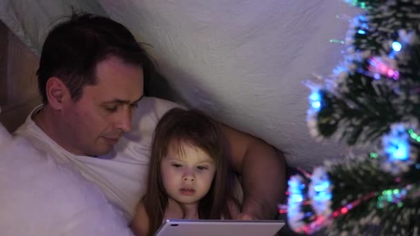 Padre e hija en la noche de Navidad, jugar y ver dibujos animados en la tableta, en una habitación para niños en una tienda de campaña con guirnaldas. bebé y padre están jugando en la habitación. concepto de infancia feliz y familia . — Vídeos de Stock