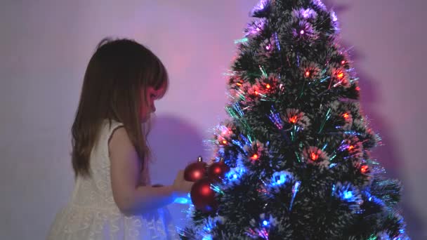 Kind versiert de kerstboom met kerstballen. klein kind speelt bij de kerstboom in de kinderkamer. dochter onderzoekt bloemenslinger op kerstboom. gelukkige kindertijd concept. — Stockvideo