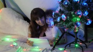 Anne ve kızı Noel akşamında, çelenklerle dolu bir çadırda, tablette çizgi film izleyip oynuyorlar. Bebek ve anne odada oynuyorlar. Mutlu çocukluk ve aile kavramı.