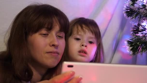 Anne ve kızı Noel akşamında, çelenklerle dolu bir çadırda, tablette çizgi film izleyip oynuyorlar. Bebek ve anne odada oynuyorlar. Mutlu çocukluk ve aile kavramı. — Stok video