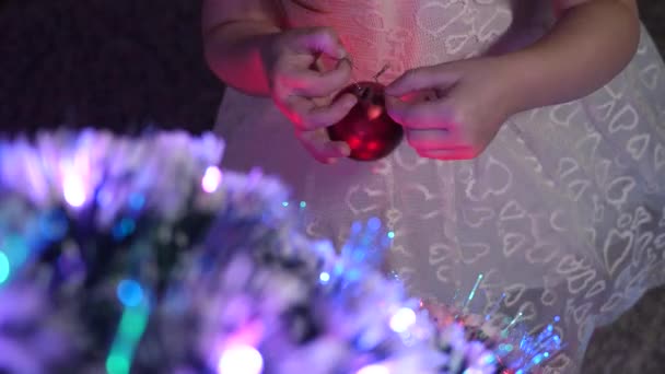 Kind versiert de kerstboom met kerstballen. klein kind speelt bij de kerstboom in de kinderkamer. dochter onderzoekt bloemenslinger op kerstboom. gelukkige kindertijd concept. — Stockvideo