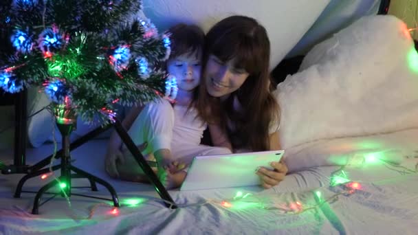 Anne ve kızı Noel akşamı çelenklerle dolu bir odada bir tablette çizgi film izliyorlar. Çocuk ve anne odada tablet bilgisayarla oynuyorlar. Mutlu çocukluk ve aile kavramı. — Stok video