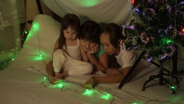 Concept van een familie Kerstmis. kinderen op kerstavond spelen op tablet, in kinderkamer op bank, in tent met bloemenslingers. zussen spelen in een kamer versierd met een veelkleurige nieuwjaarsboom — Stockvideo