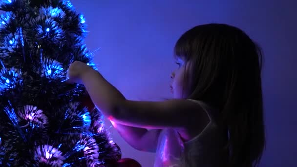 Niño decora el árbol de Navidad con bolas de Navidad. niño pequeño juega junto al árbol de Navidad en la habitación de los niños. hija examina guirnalda en el árbol de Navidad. concepto de infancia feliz . — Vídeo de stock