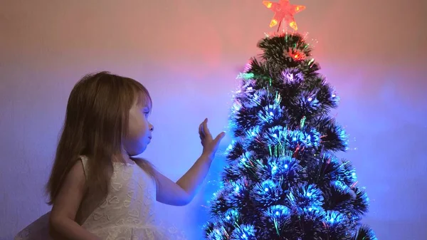 Niño pequeño juega junto al árbol de Navidad en la habitación de los niños. hija examina guirnalda en el árbol de Navidad. hermoso árbol de Navidad artificial. concepto de infancia feliz. Juegos familiares para las vacaciones de Navidad —  Fotos de Stock