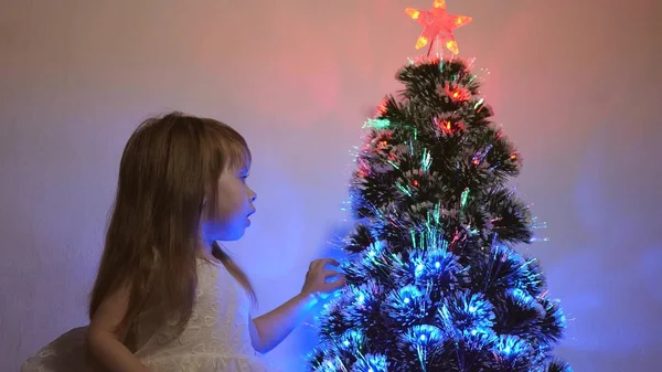 Petit enfant joue près de l'arbre de Noël dans la chambre des enfants. fille examine guirlande sur arbre de Noël. magnifique arbre de Noël artificiel. concept d'enfance heureuse. Famille joue pour les vacances de Noël — Photo