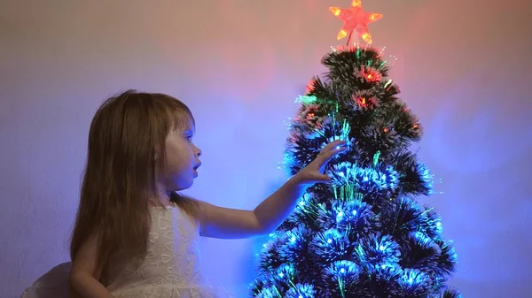 Klein kind speelt bij kerstboom in kinderkamer. dochter onderzoekt bloemenslinger op kerstboom. mooie kunstmatige kerstboom. Gelukkige kindertijd concept. Familie speelt voor kerstvakantie — Stockfoto