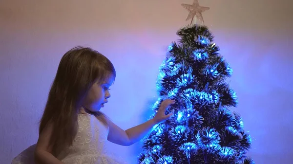 Klein kind speelt bij kerstboom in kinderkamer. dochter onderzoekt bloemenslinger op kerstboom. mooie kunstmatige kerstboom. Gelukkige kindertijd concept. Familie speelt voor kerstvakantie — Stockfoto