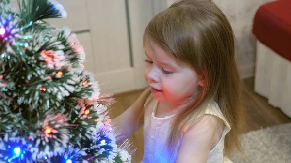 快乐的小宝宝在圣诞树旁玩耍，微笑着。 这个女孩抚摸着她的毛茸茸的美丽的云杉. 聪明的云杉 新年。 圣诞假期 — 图库照片