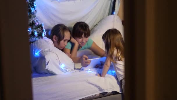 Zussen met tablet spelen in een kamer versierd met kleurrijke kerstboom. concept van een familie Kerstmis. kinderen op kerstavond spelen op tablet, in de kinderkamer op de bank slingers. — Stockvideo