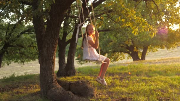 Kind swingend op een schommel in het park in de zon. jong meisje zwaaiend op touw schommel op een eiken tak. tienermeisje geniet van vliegen op swing op zomeravond in het bos. concept van gelukkig gezin en jeugd. — Stockvideo