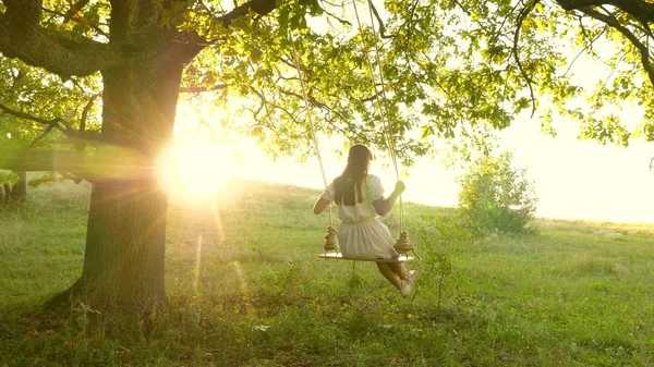太陽の下でオークの木の枝でスイングしている若い女の子空飛ぶ夢。幸せな子供時代のコンセプト。公園の白いドレスの美しい女の子。十代の女の子は森の中で夏の夜にスイングで飛行を楽しんでいます — ストック写真