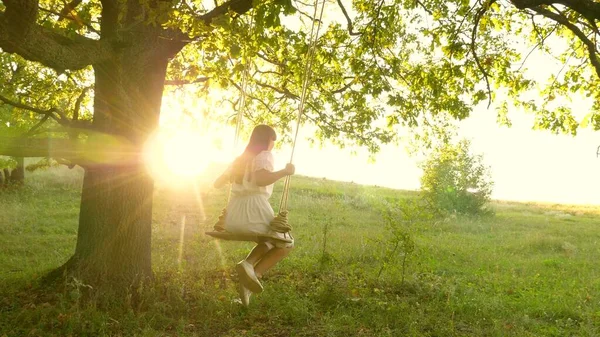 Ung flicka svänger på en gunga på en ekgren i solen. Drömmar om att flyga. Glad barndomsidé. Vacker flicka i en vit klänning i parken. tonåring flicka njuter av flygning på swing på sommaren kväll i skogen — Stockfoto