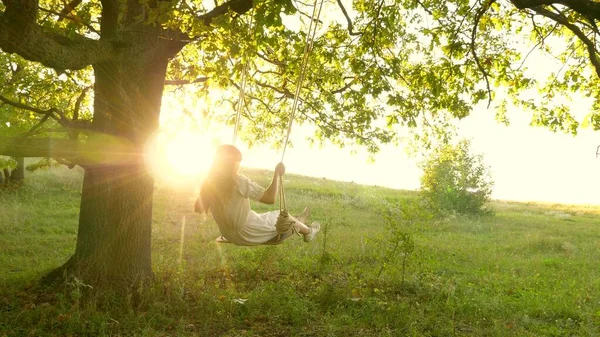 Ung flicka svänger på en gunga på en ekgren i solen. Drömmar om att flyga. Glad barndomsidé. Vacker flicka i en vit klänning i parken. tonåring flicka njuter av flygning på swing på sommaren kväll i skogen — Stockfoto