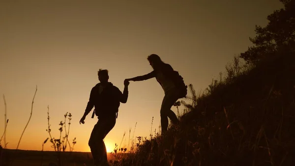 Os viajantes descem da montanha ao pôr-do-sol, de mãos dadas. conceito de aventura e viagem. trabalho de equipa de pessoas de negócios. Caminhante homem estende a mão para uma mulher viajante descendo do topo da colina . — Fotografia de Stock