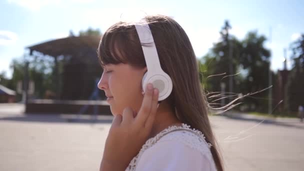 Hermosa chica viaja en una calle de la ciudad con auriculares, escucha música y sonrisas. Chica viajera pasea por la ciudad y ve los lugares de interés. En cámara lenta. adolescente salida en la ciudad . — Vídeo de stock