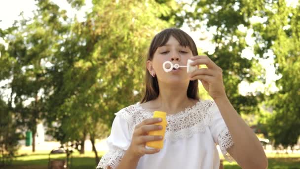 Niña jugando en el parque y soplando burbujas en la lente de la cámara. En cámara lenta. Hermosa chica soplando burbujas de jabón en el parque en primavera, verano y sonriendo . — Vídeo de stock