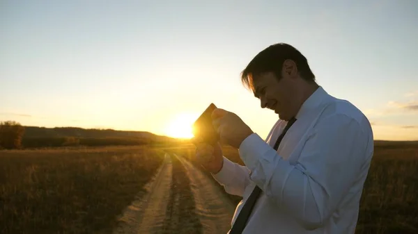 Hombre de negocios jugando juegos de tableta en el parque en los cálidos rayos de la puesta de sol. hombre jugando juegos en el teléfono inteligente. hombre feliz en camisa blanca y corbata juega en línea en la tableta . —  Fotos de Stock