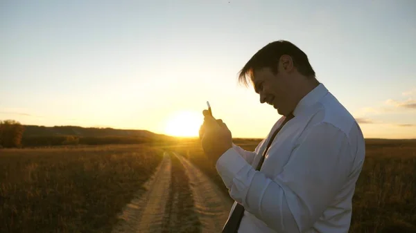 Obchodník s tabletovým zápasem v parku v teplých paprscích zapadajícího slunce. člověk hraje hry na telefonu. šťastný muž v bílé košili a kravatě hraje on-line na tabletu. — Stock fotografie