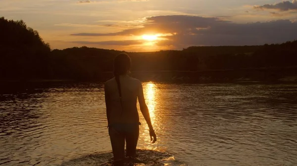 Chica entra en el agua al atardecer el sol ardiente. mujer joven se baña en la noche en el río. descansar junto al agua . — Foto de Stock