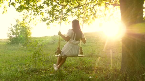 Jong meisje swingend op een schommel op een eiken tak in de zon. Dromen van vliegen. Gelukkige kindertijd concept. Mooi meisje in een witte jurk in het park. tiener meisje geniet van vliegen op swing op zomeravond in bos — Stockvideo