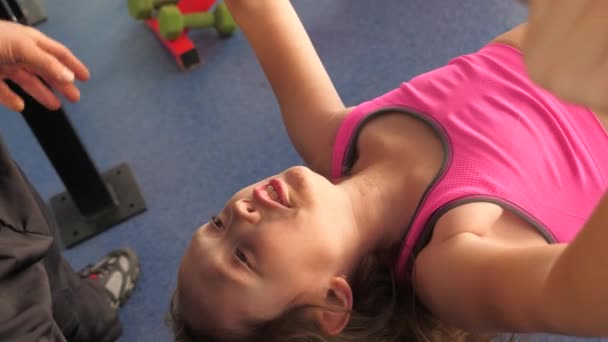 Child performs bench press lying with a trainer. girl strengthens muscles of hands. Fitness club teenager goes in for sports. Sport lifestyle concept. health promotion in gym. childrens olympic games — Stock Video