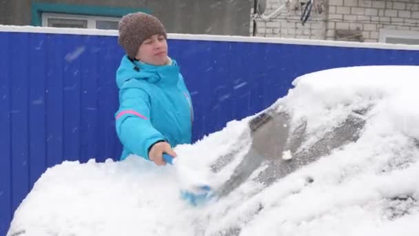 Дівчина вимітає сніг зі склянки автомобіля. Жінка очищає машину від снігу пензлем. Вишкрібання снігу та льоду з вітрового скла автомобіля. Снігопад вкрив машину . — стокове відео