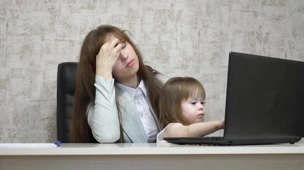 Madre stanca lavora a tavola con la sua figlioletta sul computer in ufficio. donna d'affari che lavora su un portatile con un bambino tra le braccia. Lavoro freelance femminile. Maternità moderna — Foto Stock