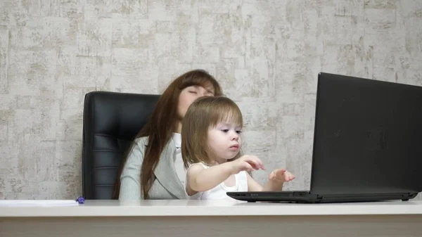 A mãe cansada trabalha à mesa com a filha no computador do escritório. Uma mulher de negócios a trabalhar num portátil com um bebé nos braços. Trabalho freelancer feminino. Maternidade moderna — Fotografia de Stock