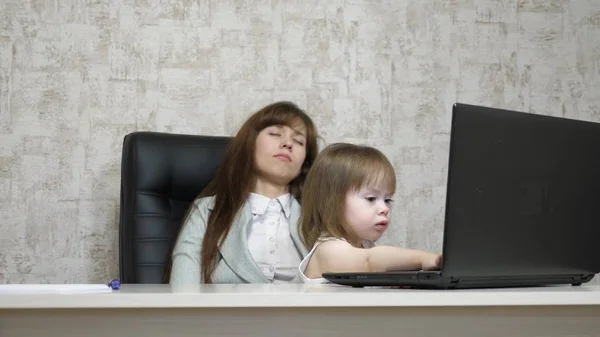 Madre cansada trabaja en la mesa con su hija pequeña en la computadora en la oficina. mujer de negocios trabajando en un portátil con un bebé en sus brazos. Trabajo independiente femenino. Maternidad moderna —  Fotos de Stock