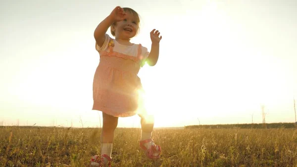 Hija pequeña va a sus padres y sonríe. En cámara lenta. niño feliz da los primeros pasos en la puesta de sol en el parque. Lindo bebé está caminando en el campo de hierba. feliz infancia y concepto familiar — Foto de Stock
