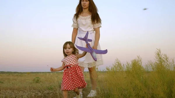 Kind mit Spielzeug spielt mit seiner Mutter auf der Wiese. Kind hält Spielzeugflugzeug in den Händen. glückliche Familie spaziert abends aus der Stadt. Tochter und Mutter gehen über das Feld. — Stockfoto