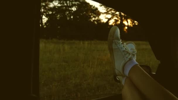 Araba camındaki bir kızın bacakları, güneş ışığı, kır yolunda araba süren bir kız. Kadın arabayla seyahat ediyor. Genç bir kadın, bacaklarını açık bir pencereden dışarı çıkararak arabayla seyahat etmeyi sever.. — Stok video