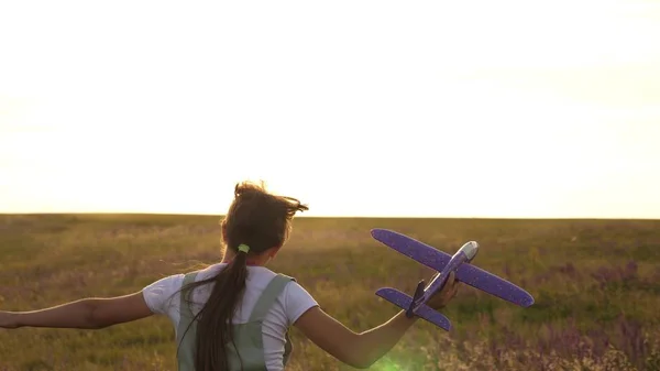 Ung flicka går med en leksak plan på fältet i strålarna av slint. barn leker leksaksflygplan. tonåring drömmer om att flyga och bli en pilot. flickan vill bli pilot och astronaut. — Stockfoto