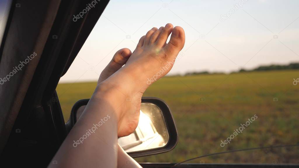 bare feet of a girl in a car window, glare of the sun, riding a car on a country road. woman travels by car. young woman likes to travel in a car, putting her legs out of an open window.