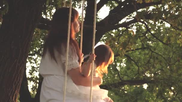 Mom shakes her daughter on swing under tree in sun. close-up. child laughs and rejoices. mother and baby ride on a rope swing on an oak branch in forest. Family fun in park, in nature. warm summer day — Stock Video