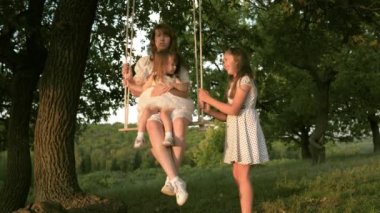 childhood and family concept. Mom shakes her daughters on a swing under tree in sun, playing with children. mother and baby ride on rope swing on an oak branch in the forest. girl laughs, rejoices.