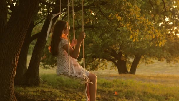 Kind swingend op een schommel in het park in de zon. jong meisje zwaaiend op touw schommel op een eiken tak. tienermeisje geniet van vliegen op swing op zomeravond in het bos. concept van gelukkig gezin en jeugd. — Stockvideo