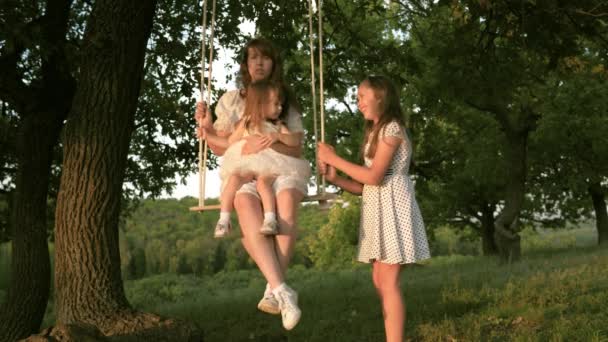 Childhood and family concept. Mom shakes her daughters on a swing under tree in sun, playing with children. mother and baby ride on rope swing on an oak branch in the forest. girl laughs, rejoices. — ストック動画