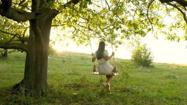 Menina livre balançando em um balanço em um galho de carvalho no sol. Sonhos de voar. Feliz conceito de infância. Menina bonita em um vestido branco no parque. menina adolescente gosta de voo no balanço na noite de verão na floresta — Fotografia de Stock
