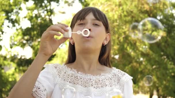 Niña jugando en el parque y soplando burbujas en la lente de la cámara. En cámara lenta. Hermosa chica soplando burbujas de jabón en el parque en primavera, verano y sonriendo . — Vídeos de Stock