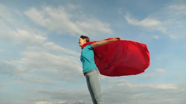 Kind speelt in rode jas, droom ervan om een held Supervrouw te worden. prachtig meisje superheld staat op het veld in een rode mantel, mantel fladderen in de wind. Langzame beweging. meisje droomt ervan om een superheld te worden. — Stockfoto