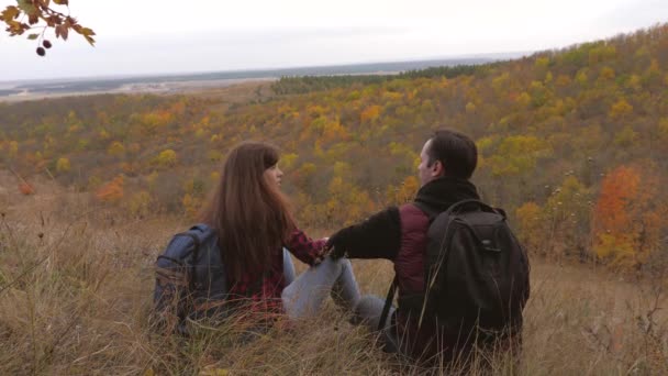 Os viajantes no outono sentam-se em uma colina e apreciam a paisagem do outono. Trabalho em equipa. O conceito de turismo desportivo. turistas homem e mulher estão descansando e olhando para a distância do topo da montanha . — Vídeo de Stock