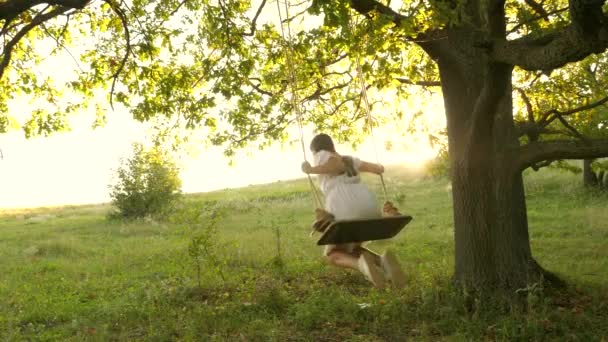 비행의 꿈. 십 대 소녀들은 숲 속에서 여름 저녁에 비행기를 타는 것을 좋아 합니다. 태양에 있는 오크 가지 위에서 그네를 타는 자유 소녀. 행복 한 어린 시절의 컨셉. 공원에서 하얀 드레스를 입은 아름다운 소녀. — 비디오