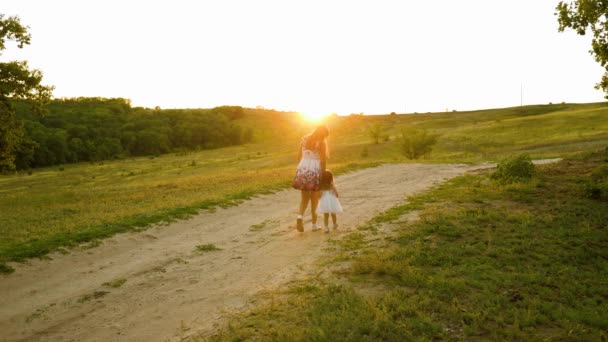 Mutlu aile kavramı. Anne ve bebek el ele tutuşup akşam vakti güzel bir gün batımı ışınları altında parkta yürürler. Beyaz elbiseli küçük kız annesiyle birlikte yolda yürüyor. Mutlu aile seyahatleri — Stok video