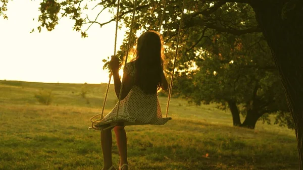 Bambino che oscilla su un'altalena nel parco al sole. giovane ragazza che oscilla su un'altalena di corda su un ramo di quercia. teen girl gode di volo su altalena la sera d'estate nella foresta. concetto di famiglia felice e infanzia. — Foto Stock