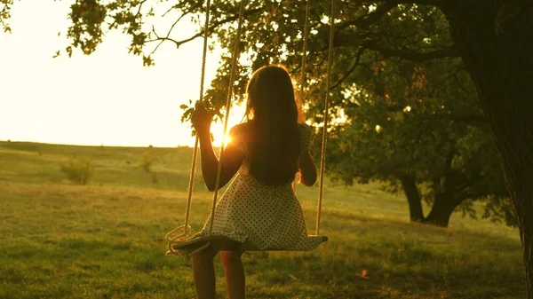 Bambino che oscilla su un'altalena nel parco al sole. giovane ragazza che oscilla su un'altalena di corda su un ramo di quercia. teen girl gode di volo su altalena la sera d'estate nella foresta. concetto di famiglia felice e infanzia. — Foto Stock