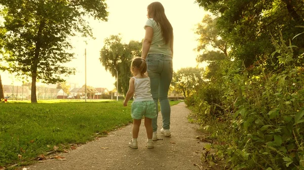 Маленькая дочь и мама идут по тропинке, держась за руки. Ребенок держит маму за руку. Мать и ребенок отдыхают в парке. Понятие счастливого детства. Счастливая семья с ребенком гуляет вечером за городом . — стоковое фото