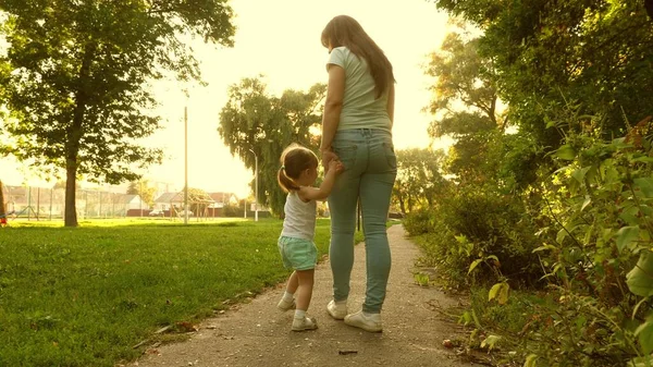 little daughter and mom walk path holding hands. baby holds moms hand. Mother and baby rest in park. The concept of a happy childhood. A happy family with a child walks in evening out of town.