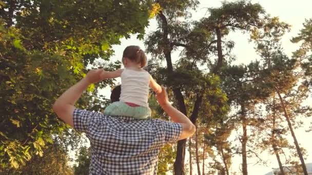 Apa a szeretett gyermeke vállát hordozza a parkban. Apa a lányával a vállán sétál a fák alatt. A gyerek a szüleivel szabadnapos. A boldog család a parkban pihen.. — Stock videók